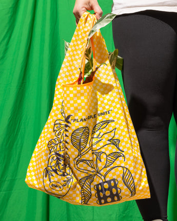 A person holding a yellow checkered reusable bag with plant illustrations and text on it.