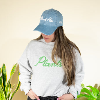 A person wearing a blue denim baseball hat with the text 'Plant Hoe' on it and a gray sweatshirt with the word 'Plants' written across the front in green lettering.