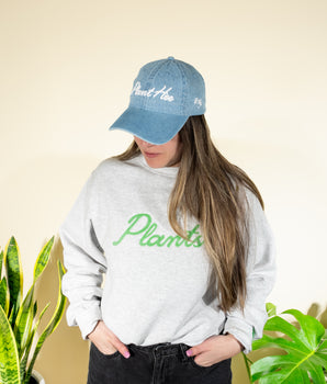 A person wearing a blue denim baseball hat with the text 'Plant Hoe' on it and a gray sweatshirt with the word 'Plants' written across the front in green lettering.