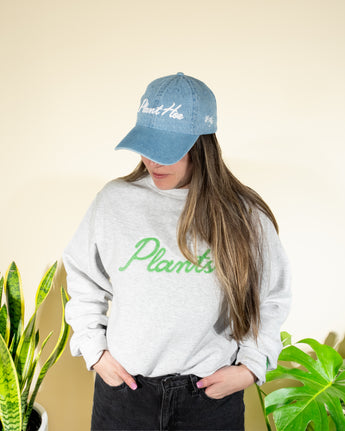 A person wearing a blue denim baseball hat with the text 'Plant Hoe' on it and a gray sweatshirt with the word 'Plants' written across the front in green lettering.