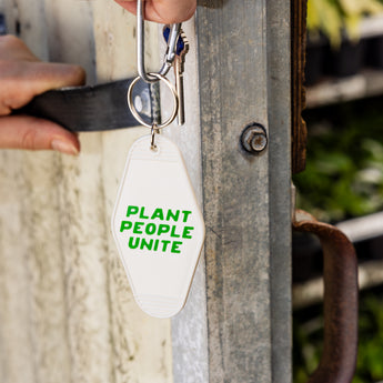 A white keychain with green text that reads 'Plant People Unite' attached to a keyring, held up against a metallic background.