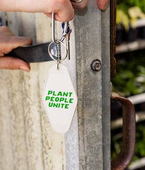 A white keychain with green text that reads 'Plant People Unite' attached to a keyring, held up against a metallic background.