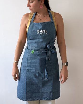 Person wearing a blue denim apron with 'BWH Plant Co' logo against a plain background