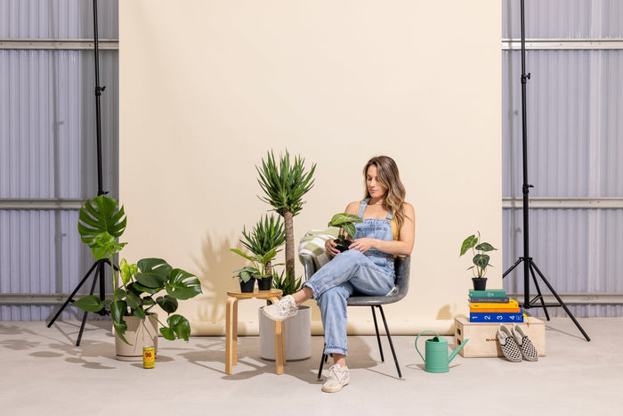 A person in overalls sitting on a chair surrounded by plants, a watering can, and books, against a light beige background in a studio setting.