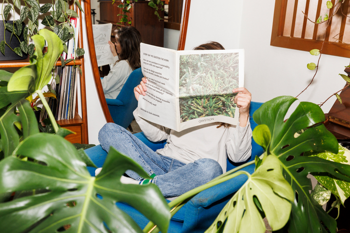 A person sits cross-legged in a blue chair, reading a magazine, surrounded by lush indoor plants.