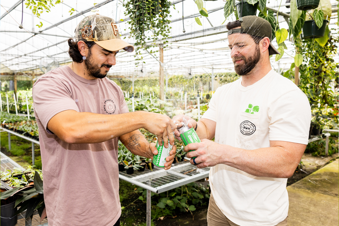 Two men in a greenhouse are clinking green canned beverages, with spray splashing from the cans.