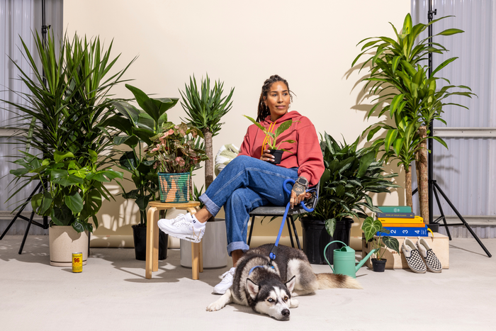 A woman in a red hoodie holds a potted plant, sitting among lush greenery with a Husky lying at her feet.