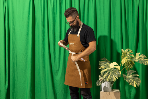 A man in a brown apron ties the waist straps, standing near a variegated Monstera plant with a green backdrop.