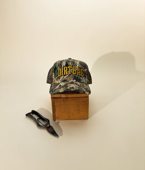Camouflage cap with gold 'DIRTBAG' text across front. Placed on a wooden block on beige background. 